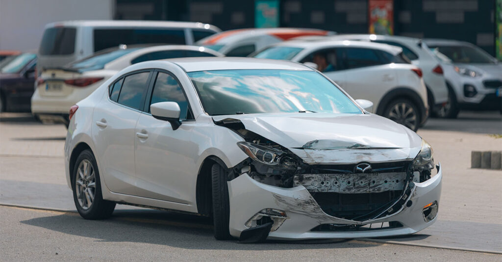 Small white Mazda hatchback which front smashed in and bumper hanging off